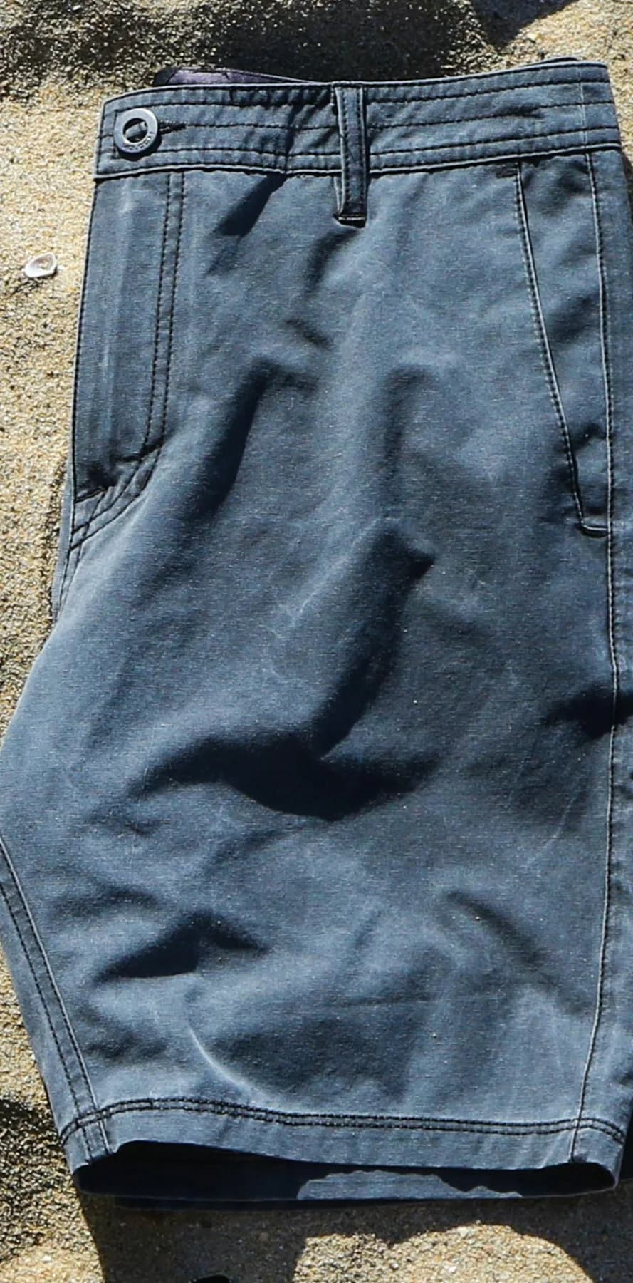 Board short cinza inteiro em flat lay sobre areia, com cós e barra visíveis